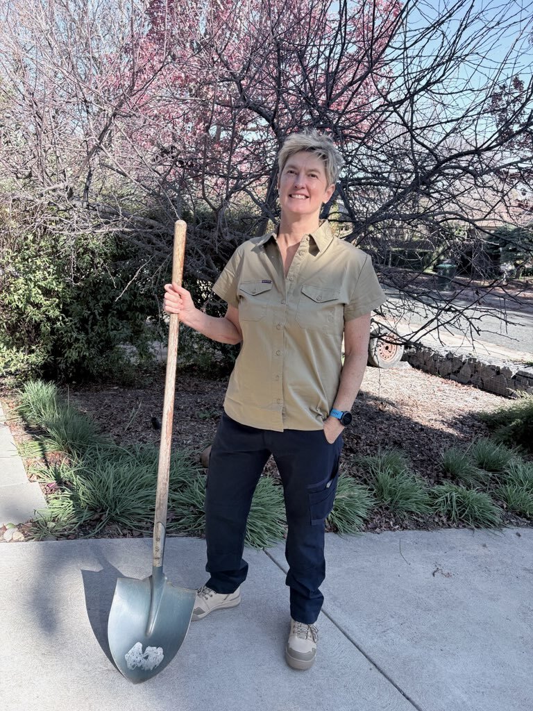 Sue standing in a garden holding a long-handled digging shovel, smiling, with a blossoming tree behind her